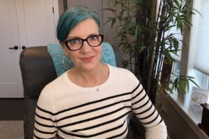Pretty white woman with teal-colored hair, a striped swearing, large black frame glasses.Sitting in an office chair